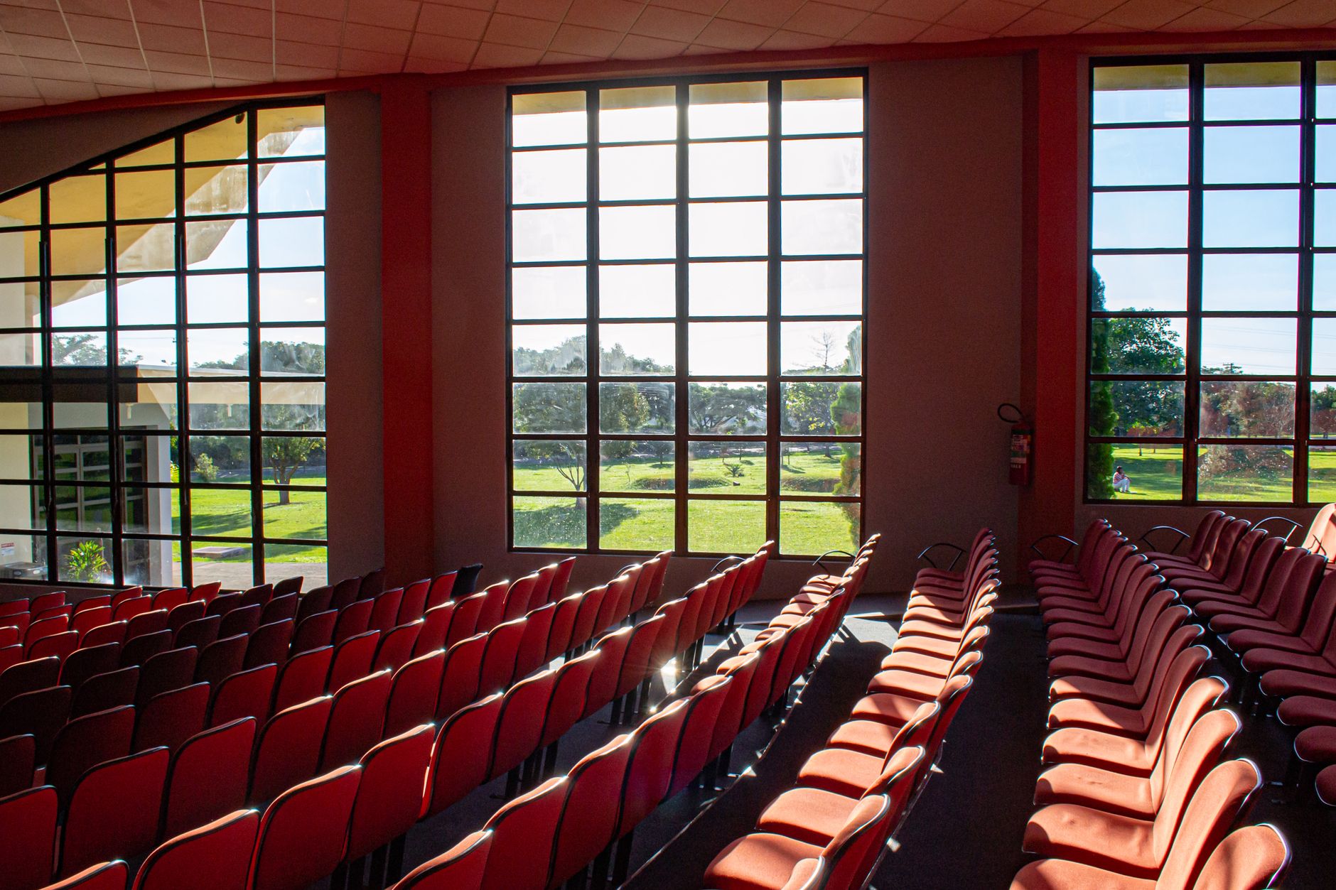 Bright auditorium with red chairs, large windows, and a sunny outdoor view.