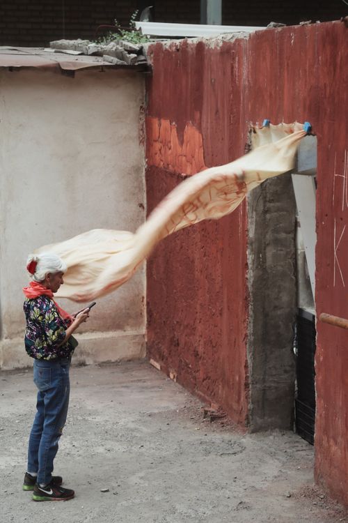 Free Elderly woman standing outdoors, scarf dancing in the wind against rustic red wall. Stock Photo