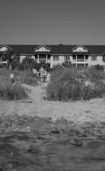 Black and white photo of a seaside hotel with two adults walking by the beach.