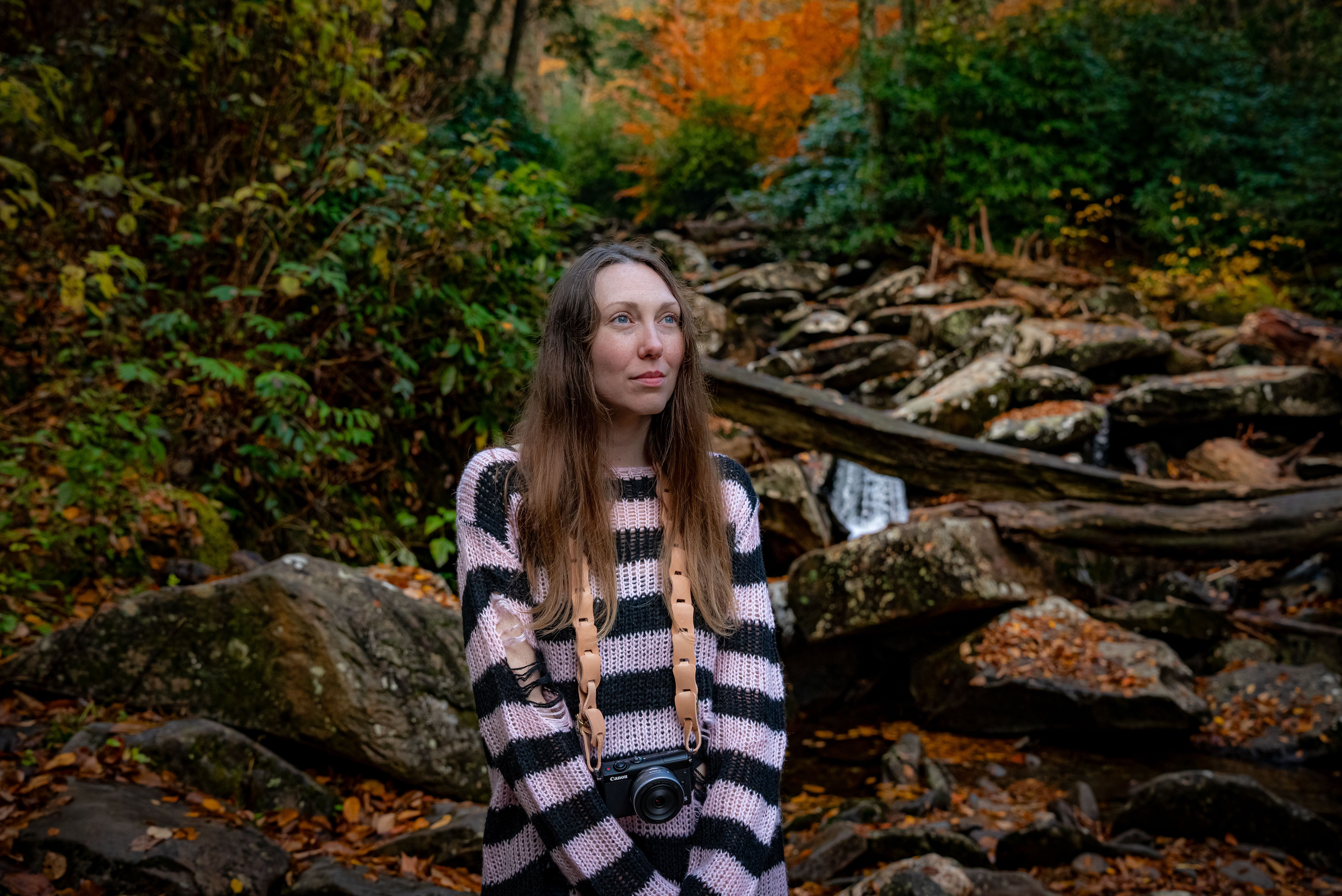 A woman in a striped sweater explores a vibrant autumn forest with her camera.