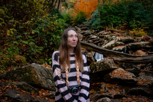 A woman in a striped sweater explores a vibrant autumn forest with her camera.