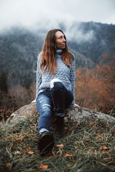 A woman in a cozy sweater sits on a rock surrounded by an autumn mountain landscape.