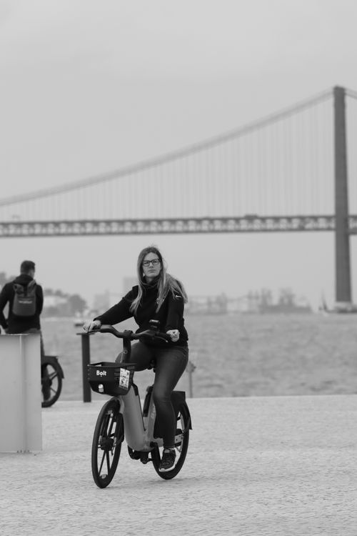 Free Black and white photo of a woman cycling by the 25 de Abril Bridge in Lisbon, Portugal. Stock Photo