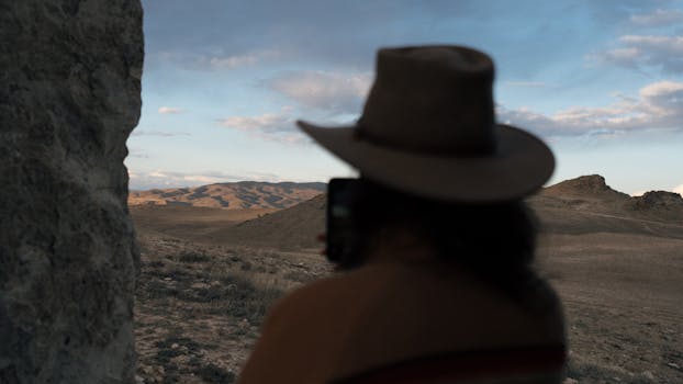 A cowboy in a hat views a sprawling desert landscape under a serene sky.