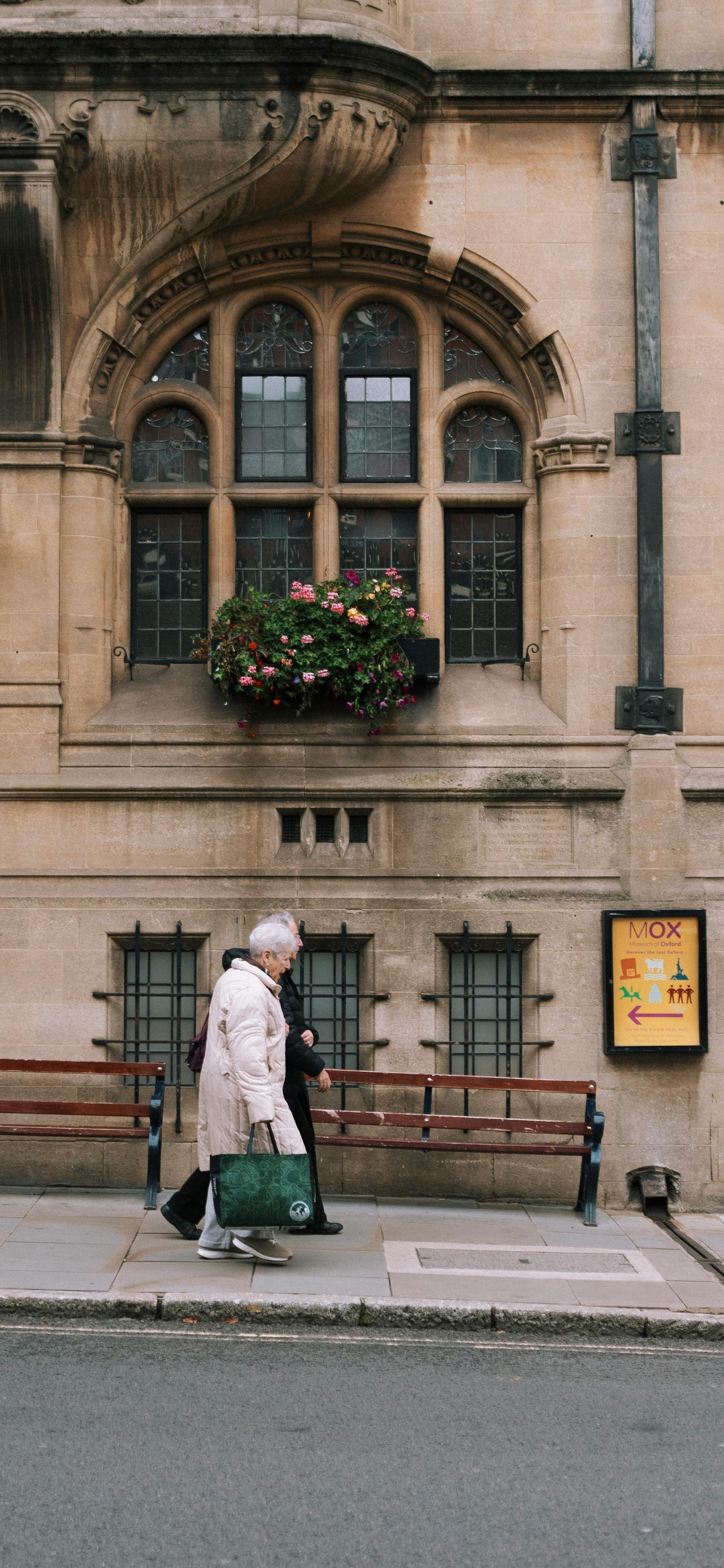 An elderly couple walks past a historic building with ornate architecture and flower boxes.