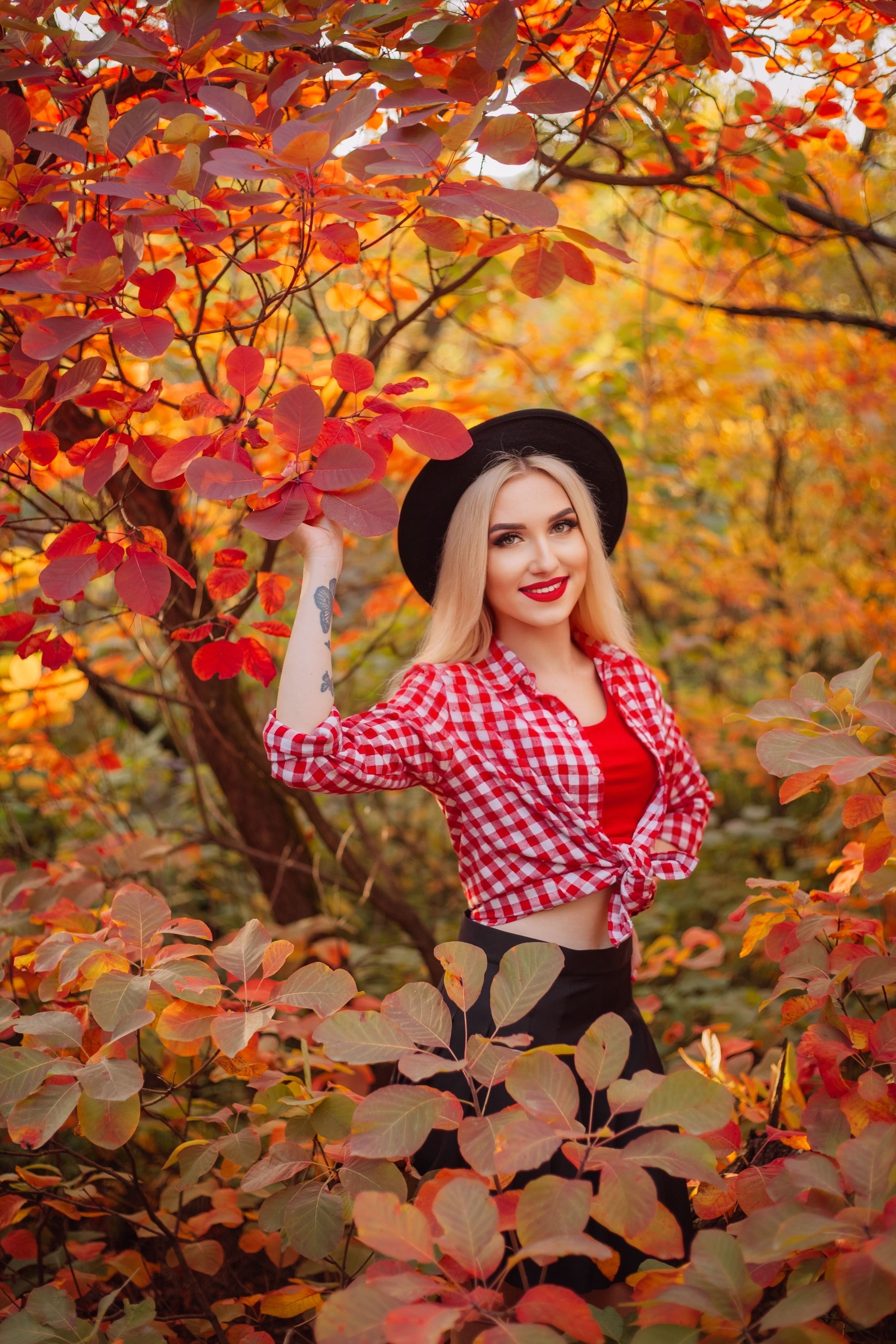 Photograph of a smiling woman in a red checkered shirt amidst vibrant autumn foliage.