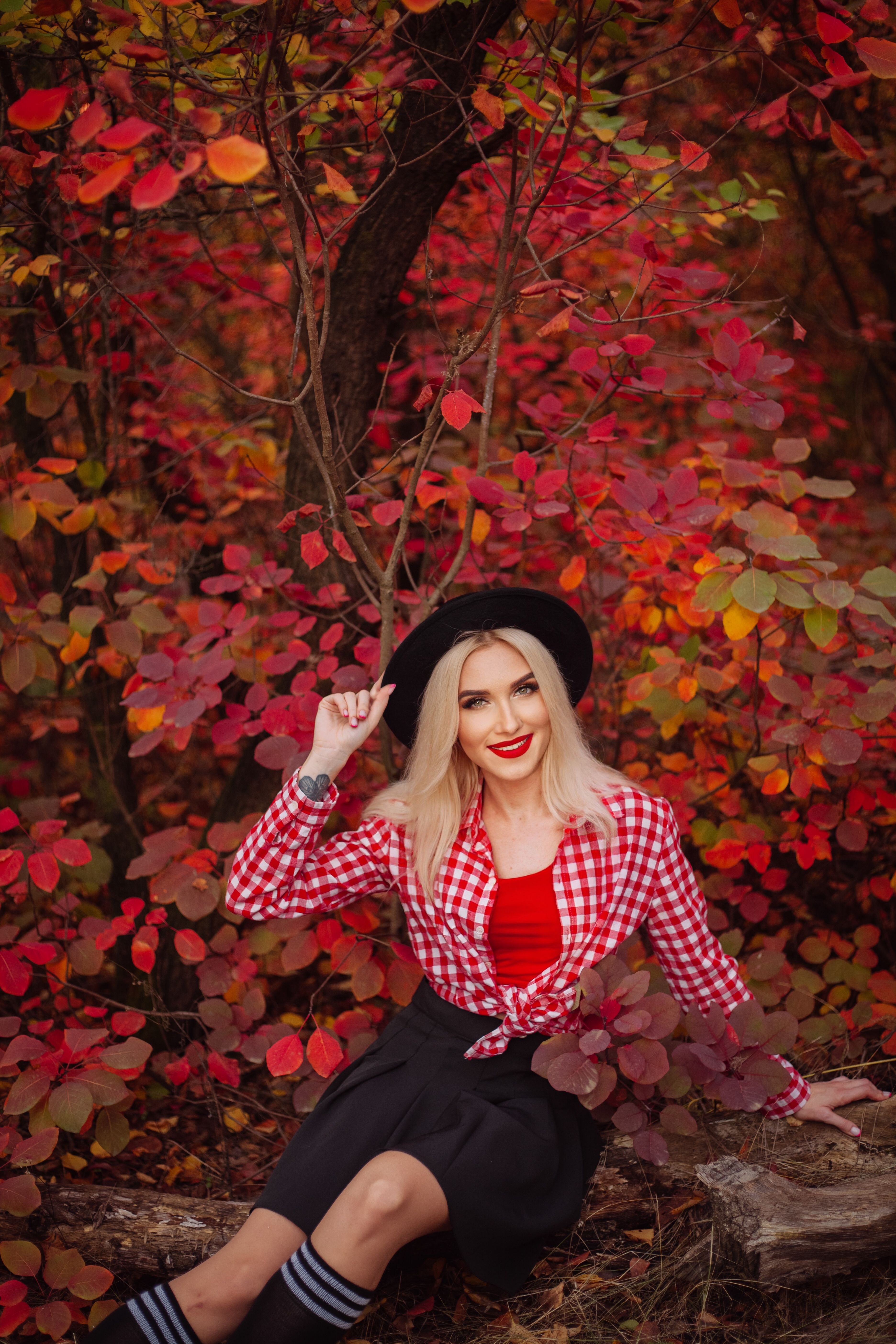 Blonde woman in red plaid shirt and hat smiling amidst vibrant autumn foliage.