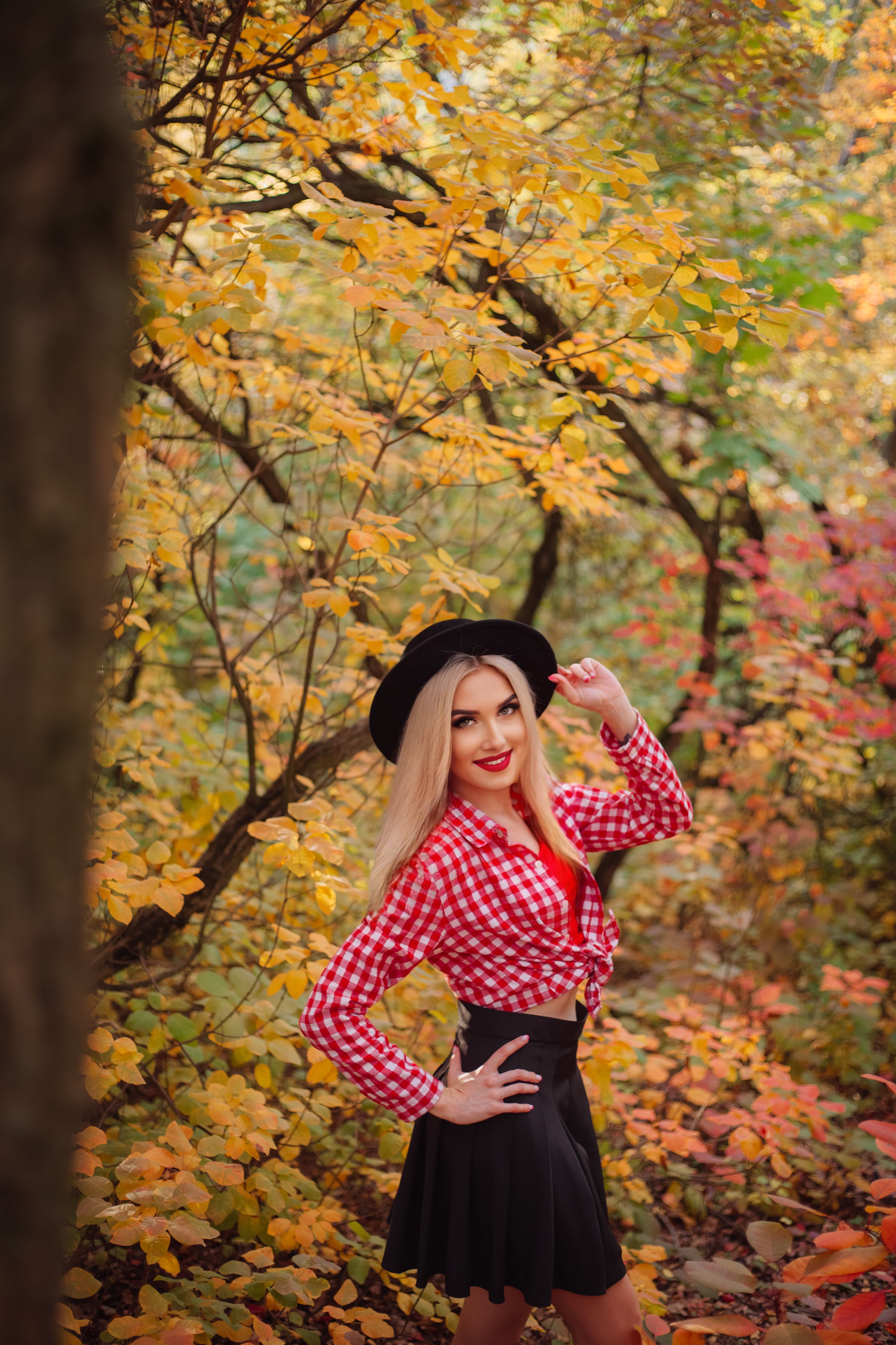 A cheerful woman in a red checkered shirt poses in a vibrant autumn forest setting.