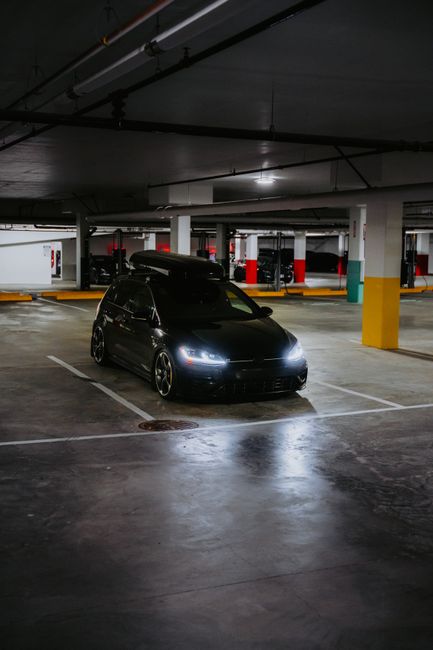 A stylish black car parked in a dim indoor parking garage with headlights on.