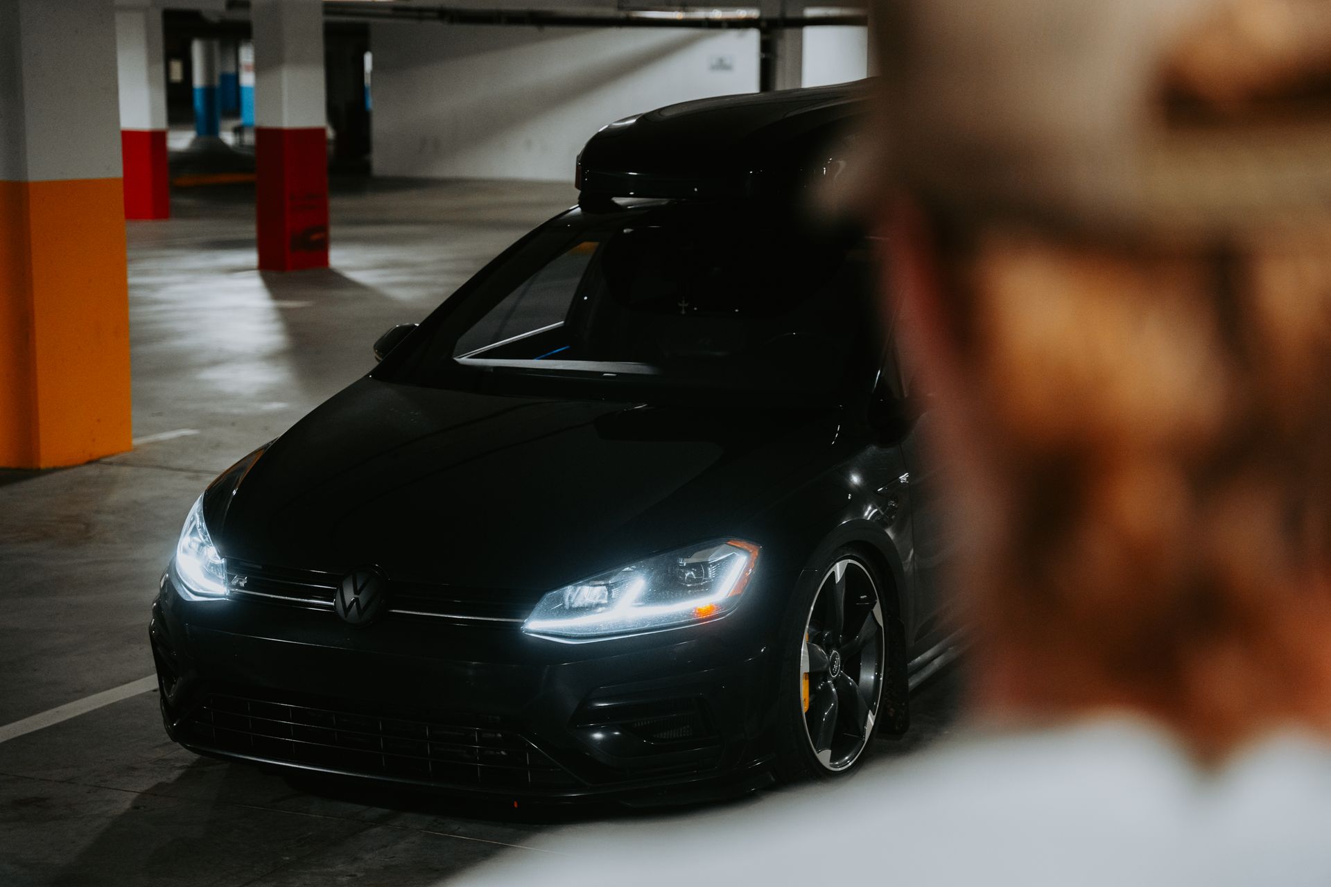 A sleek black car with headlights on inside an underground parking garage.
