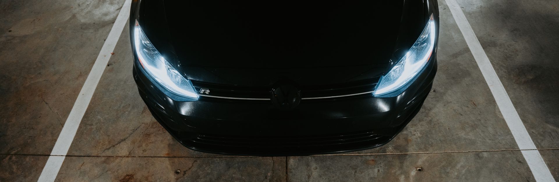 Top view of a sleek black car with headlights on, parked in an underground garage.