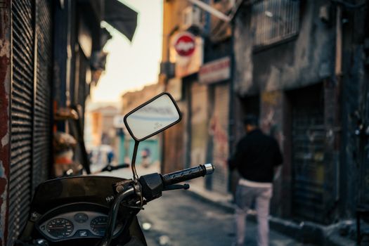 A motorcycle parked in a dimly lit urban alley with a blurred pedestrian in the background.