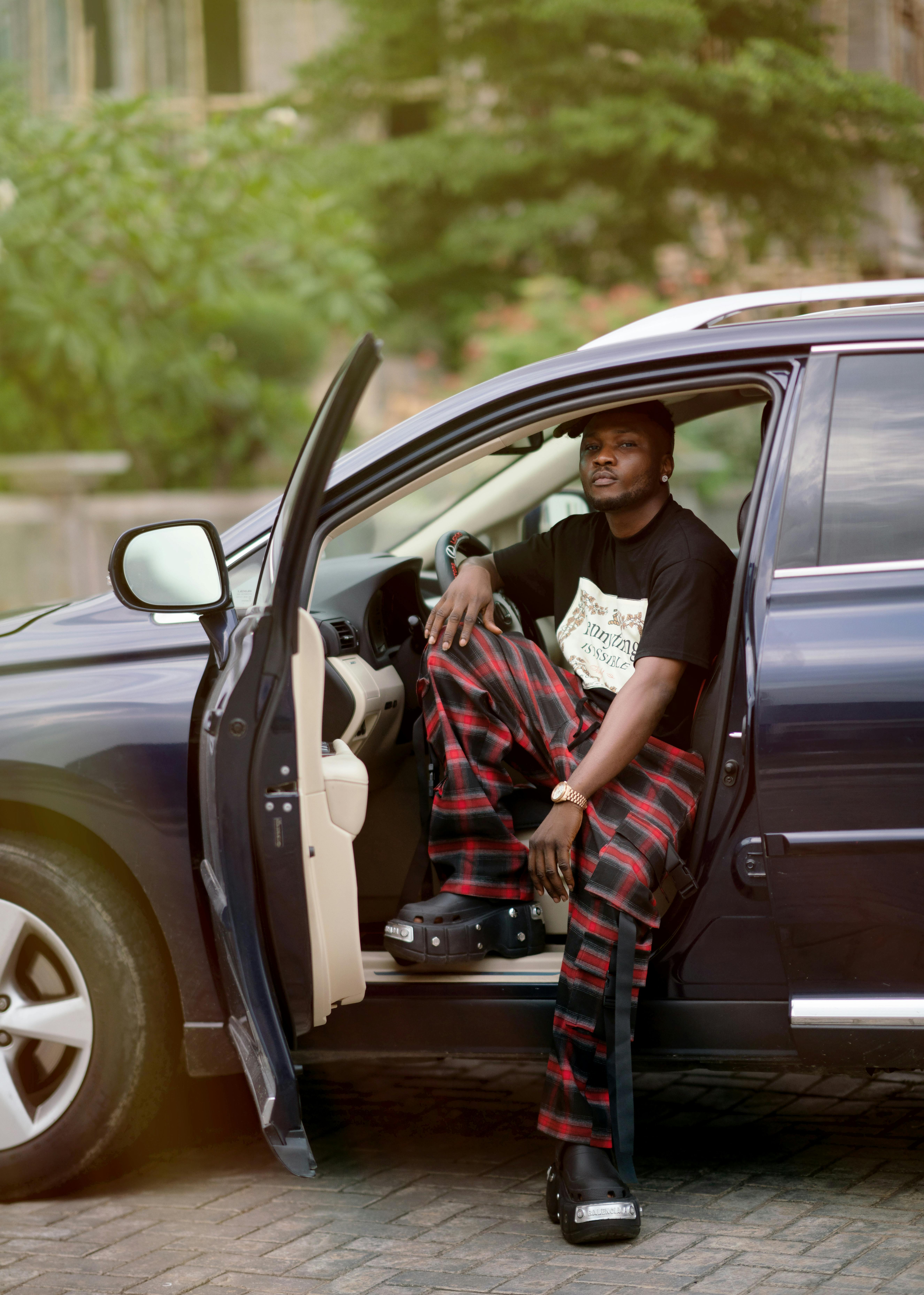 Man in plaid pants sitting in an open car door outdoors.