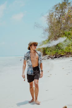 Man wearing summer attire walks on a sandy beach with lush greenery under clear sky.