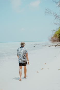 A man in a hat walks on a serene tropical beach, leaving footprints on the sand.