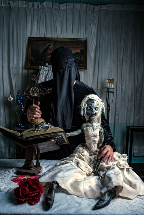 Free A veiled figure holding a cross and a doll in a dimly lit room setting, evoking mystery and intrigue. Stock Photo