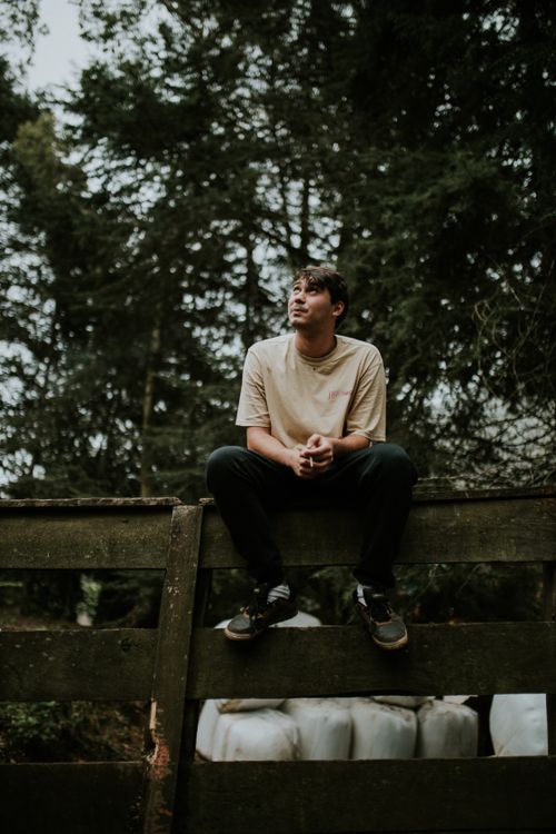Free Casual young man sitting on a wooden fence in a serene forest setting, contemplating nature. Stock Photo