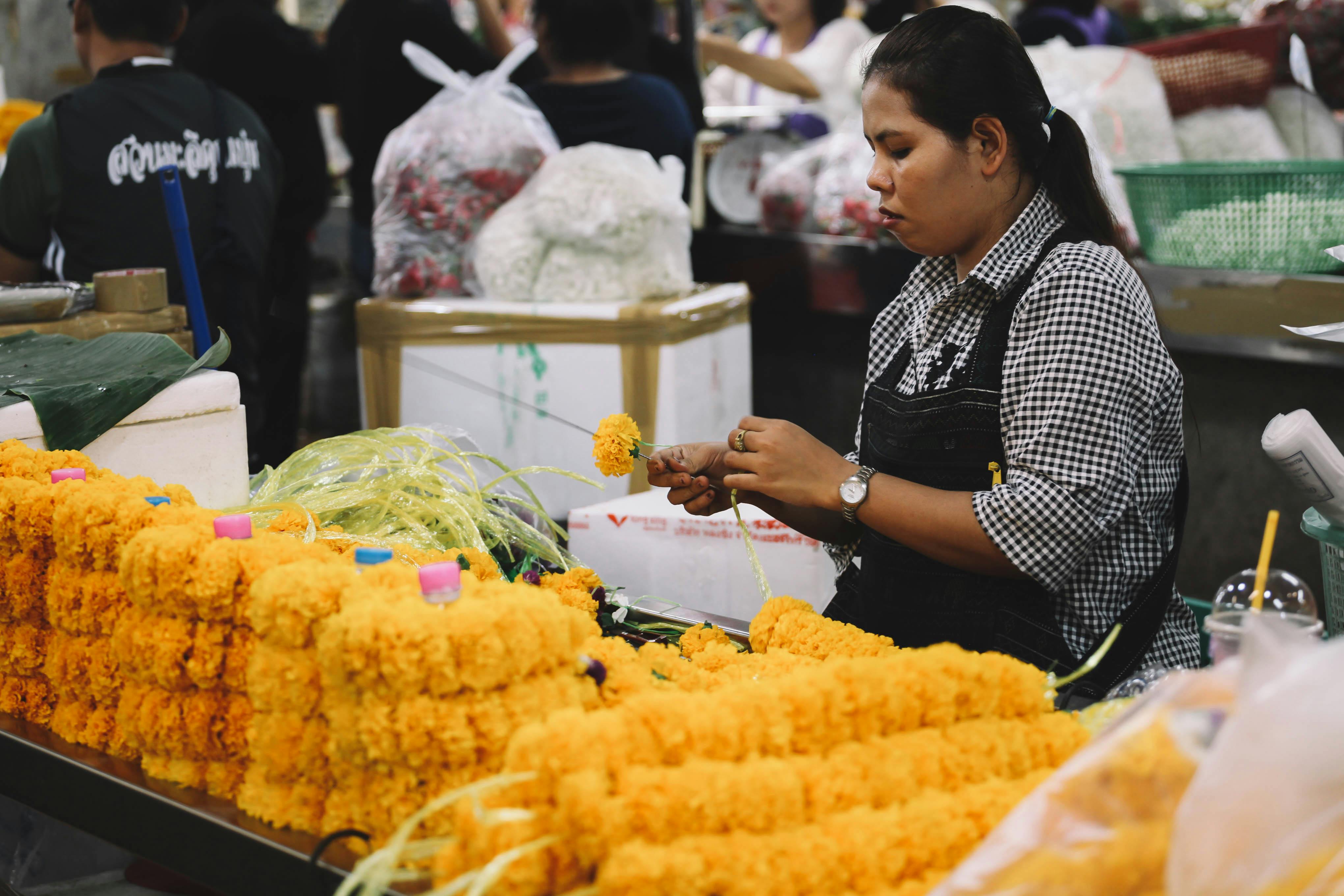 Woman handcrafting vibrant marigold garlands in a bustling Bangkok market scene. - Colabora Birmania