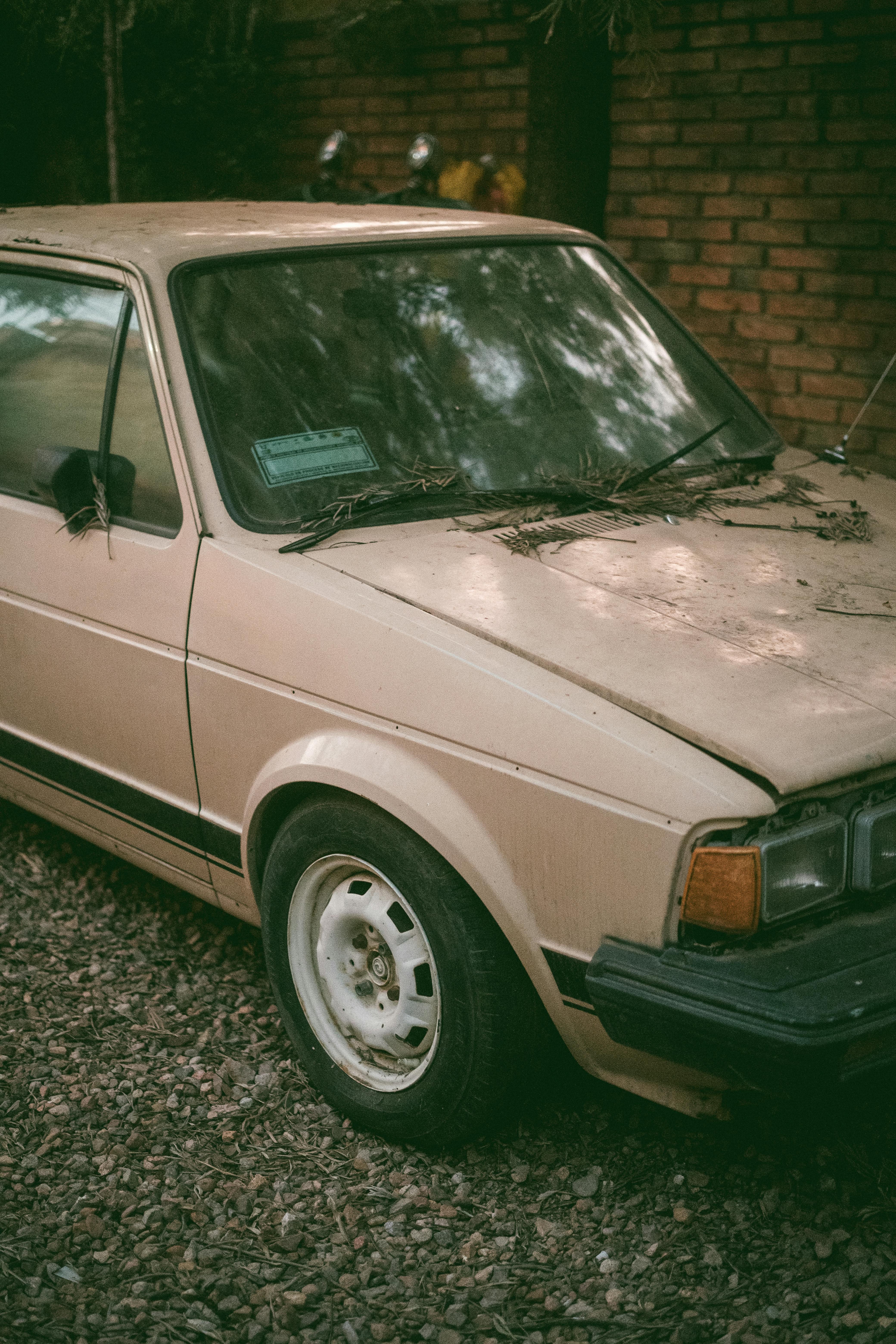 Old beige car parked outdoors on gravel, surrounded by rustic brick and overgrown foliage.