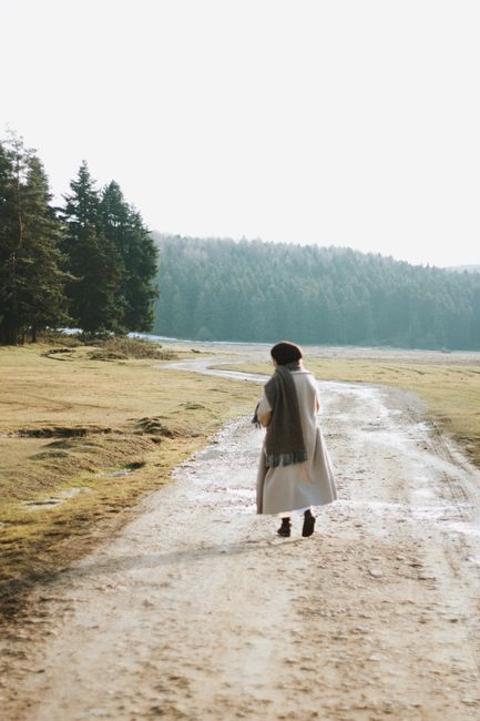 A woman in a coat walks along a rural dirt path amidst scenic countryside with tall trees.