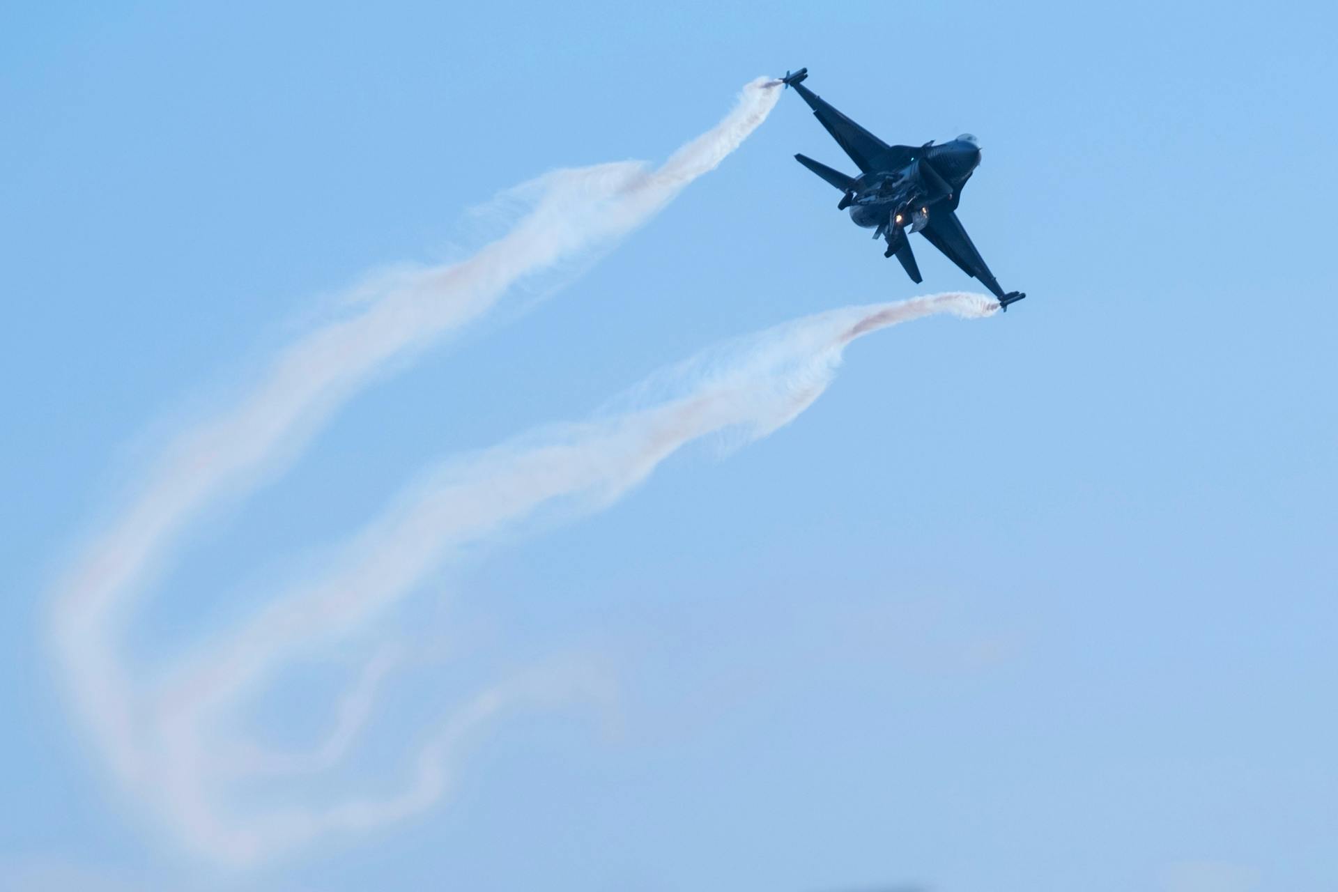 Fighter jet performing an agile maneuver in clear blue sky, leaving vapor trails.