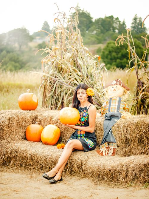 Free A woman in colorful attire poses with pumpkins and a scarecrow on a hay bale outdoors. Stock Photo