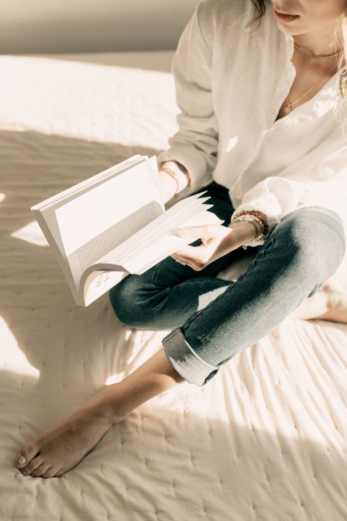 Free A woman in casual attire reading a book on a sunlit bed, creating a cozy and relaxed atmosphere. Stock Photo