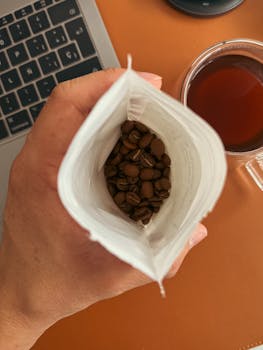 Fresh coffee beans in a white bag with a laptop and coffee cup nearby, perfect for work-from-home concept.