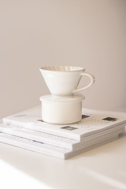 Elegant minimalist setup featuring a white coffee dripper atop stacked magazines on a bright surface.