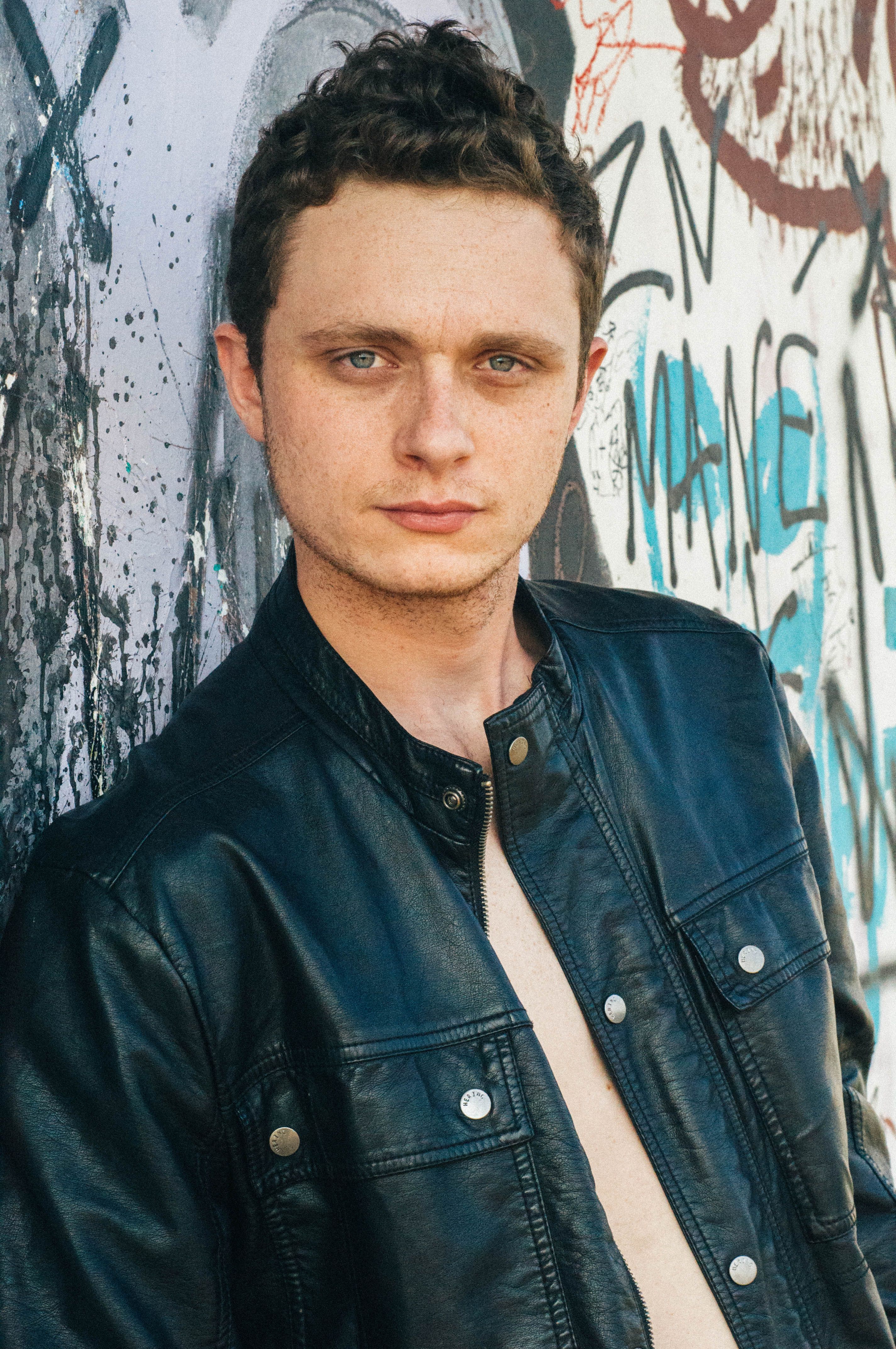 Portrait of a man in a leather jacket standing against a graffiti wall outdoors.