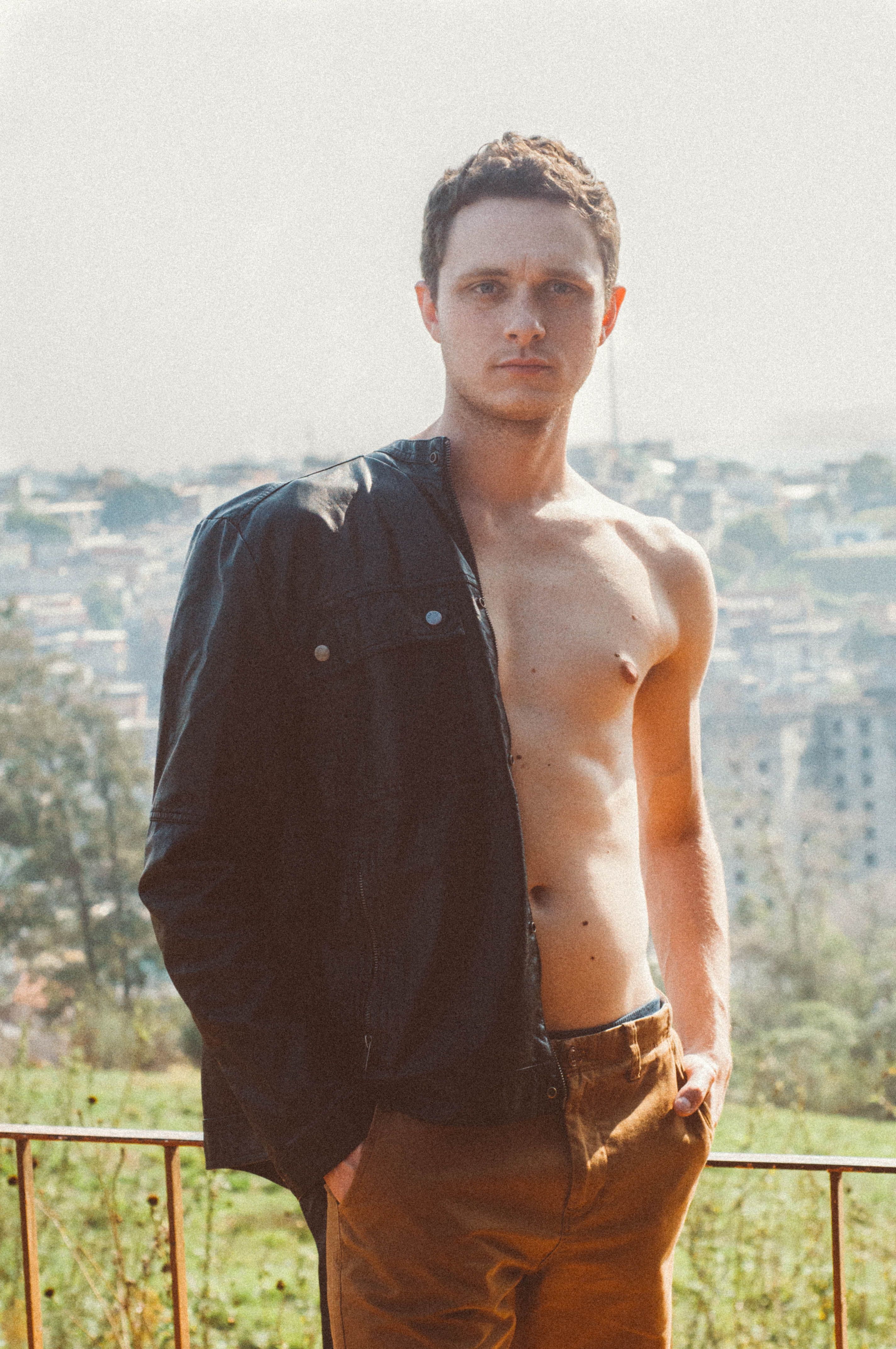 Young man in half-dressed leather jacket posing outdoors with cityscape backdrop.