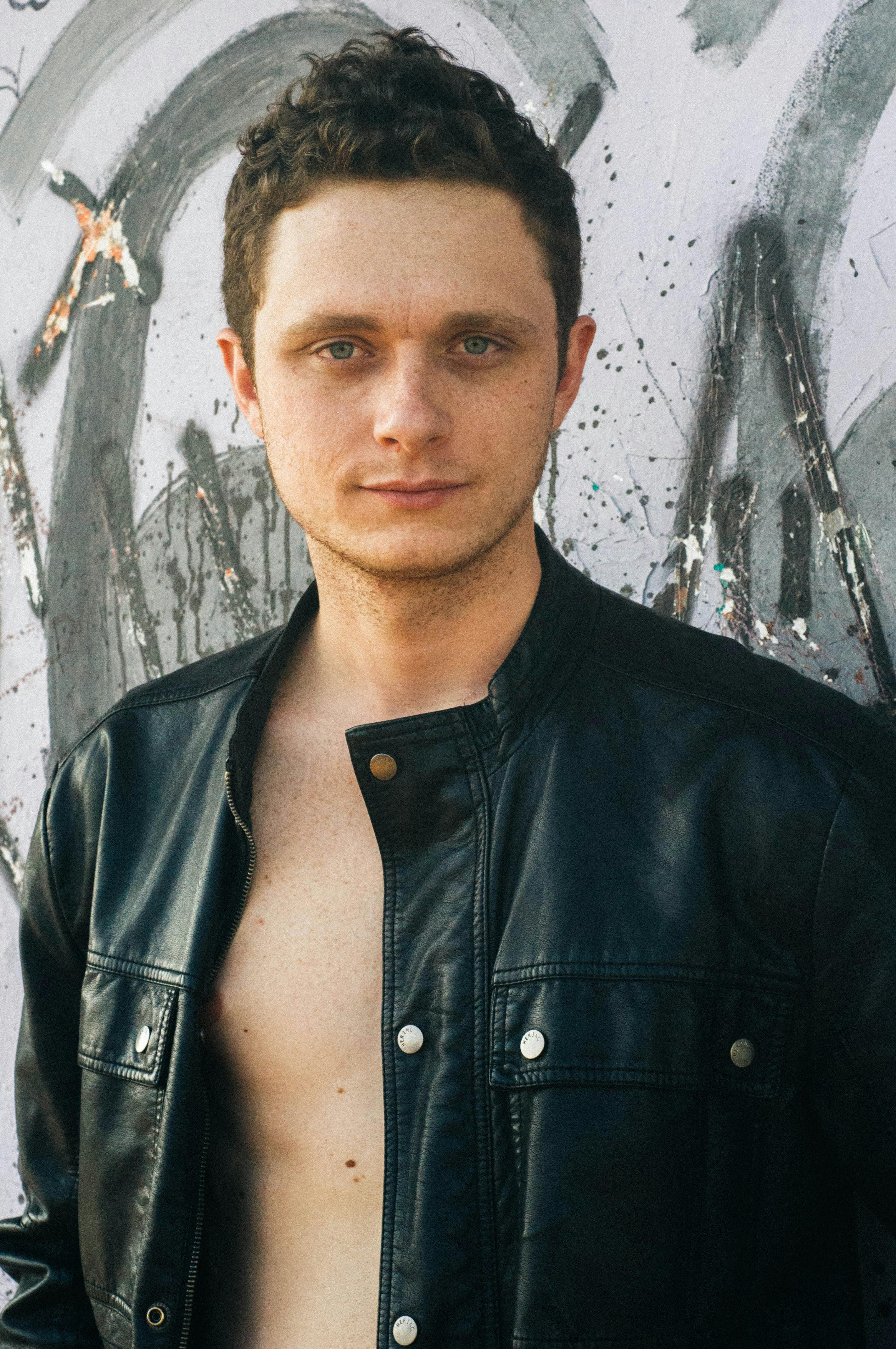 Young man posing outdoors in leather jacket against graffiti wall.