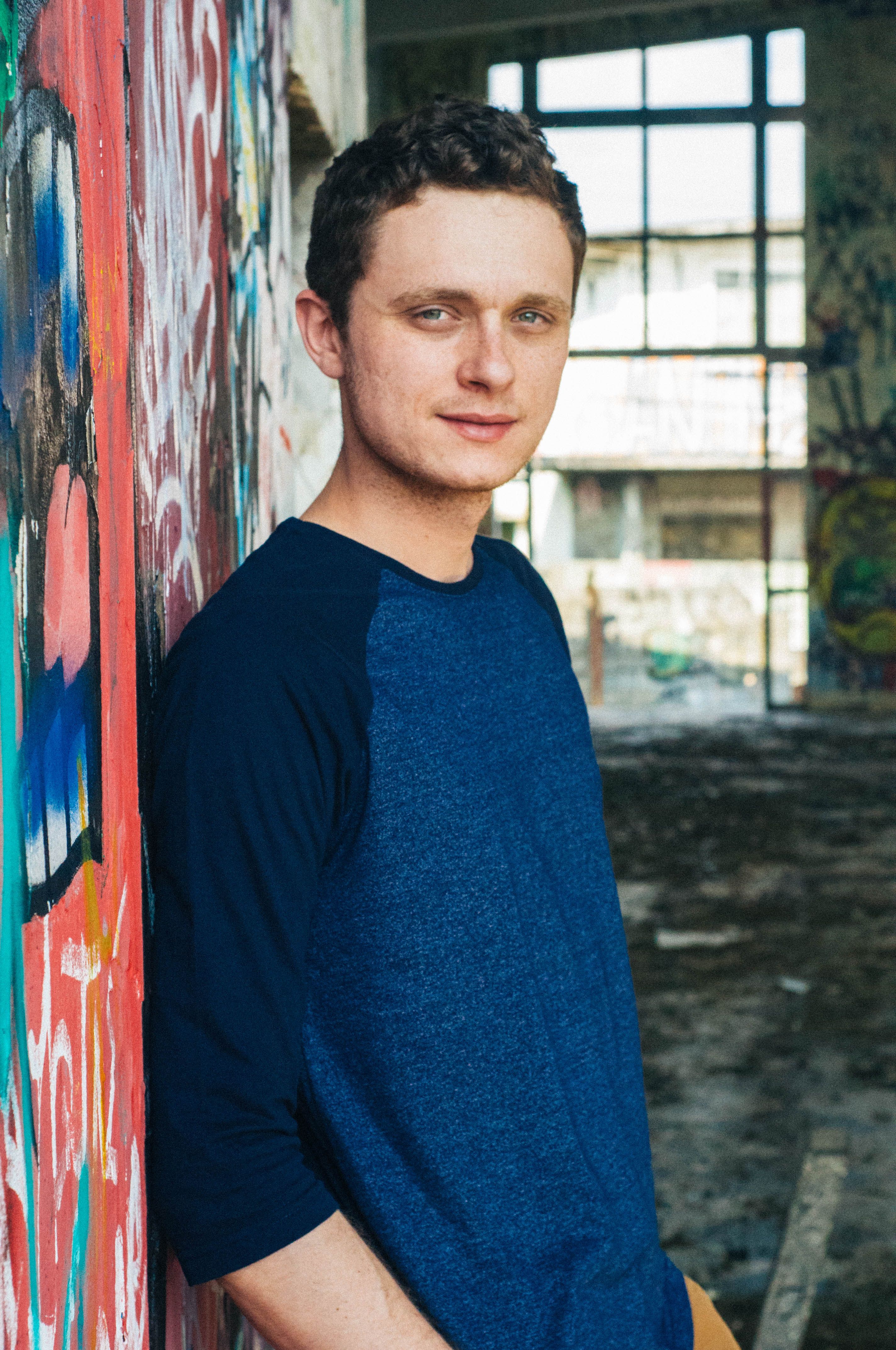 Young man in casual attire posing confidently against a graffiti-covered wall in an abandoned building.