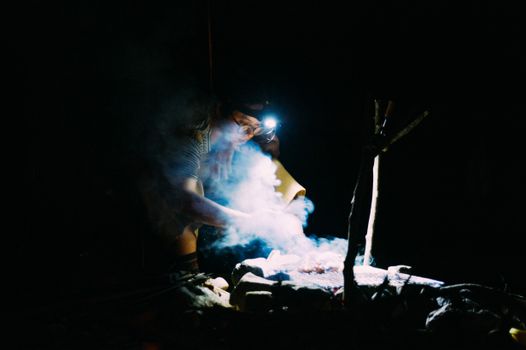 A person cooking at night over a smoky campfire with a headlamp.