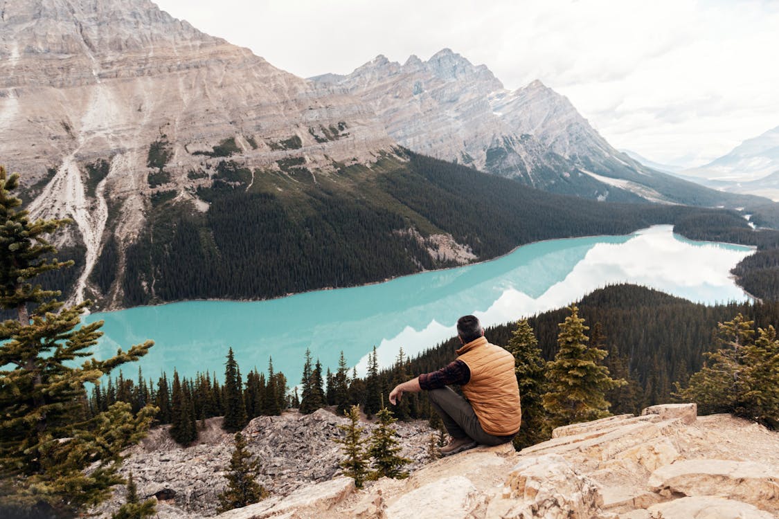 Man Wearing Brown Vest Sitting on Mountain Looking at Lake