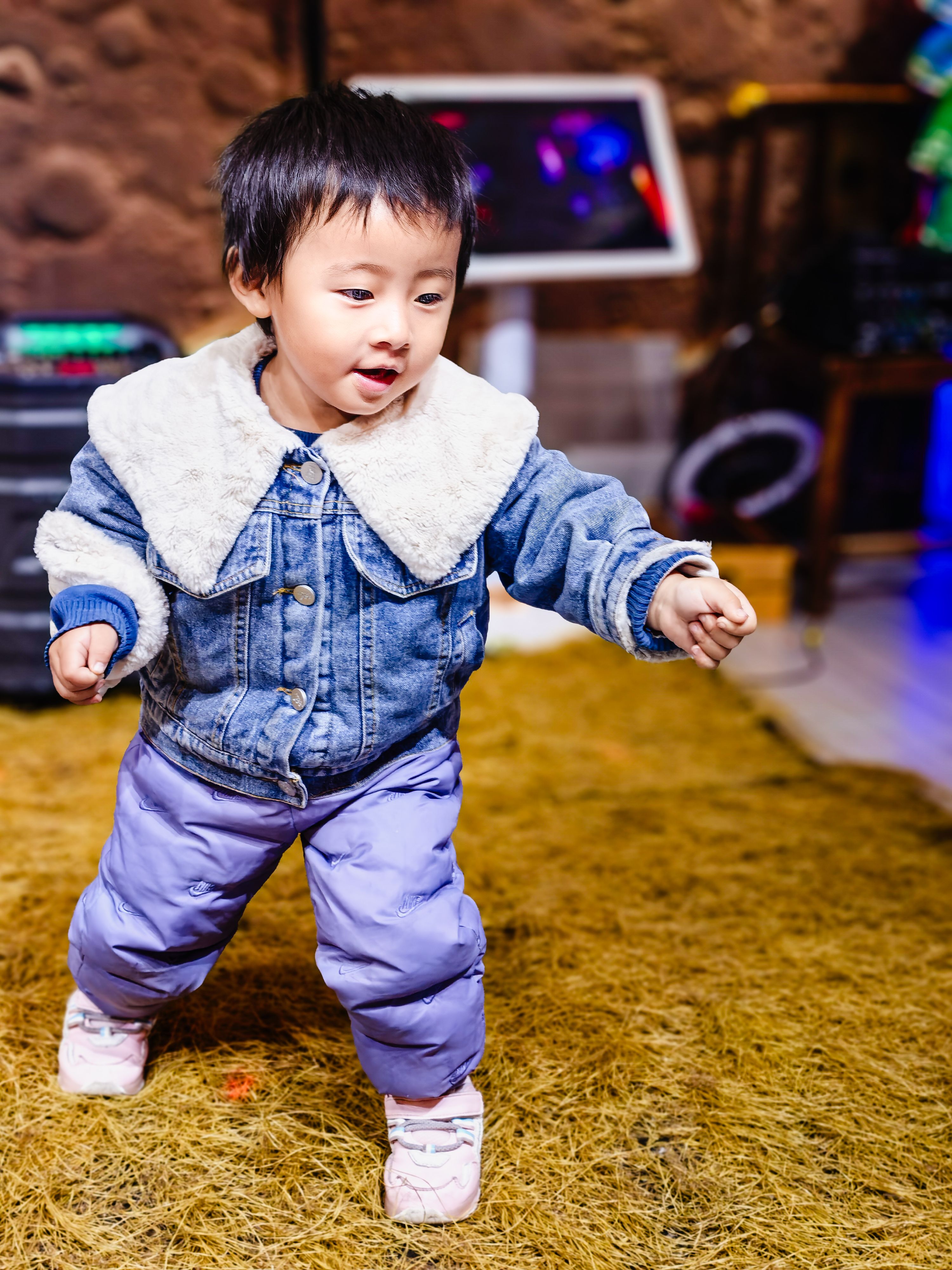 Charming toddler in a stylish denim jacket and blue pants exploring an indoor play area.
