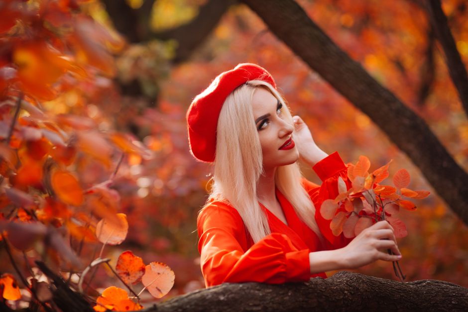 Blonde woman in red beret surrounded by vibrant autumn leaves in a forest setting.