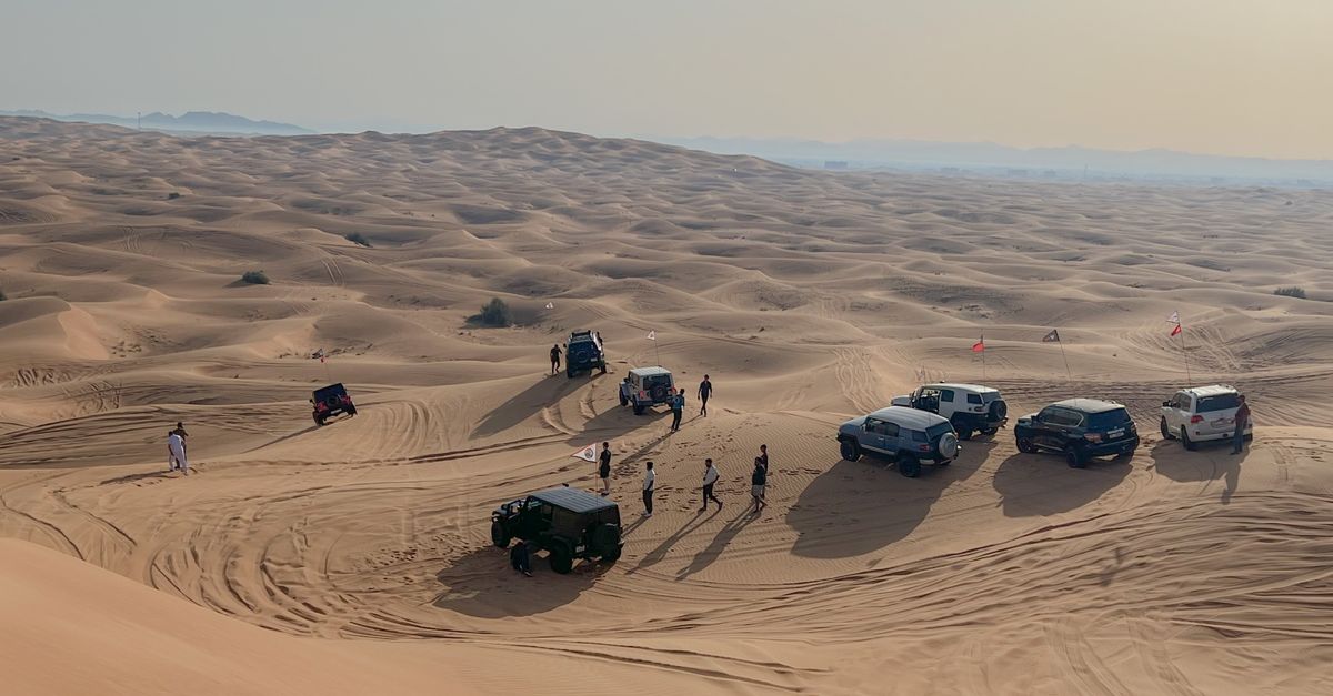 A white sedan parked on a roadside in the desert with its hood open and smoke billowing from the engine.