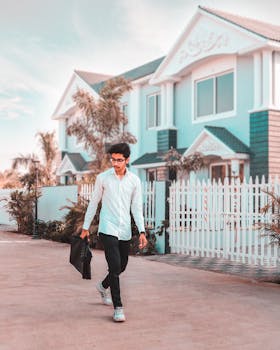 A fashionable young man in casual attire walks past a modern house on a sunny day.
