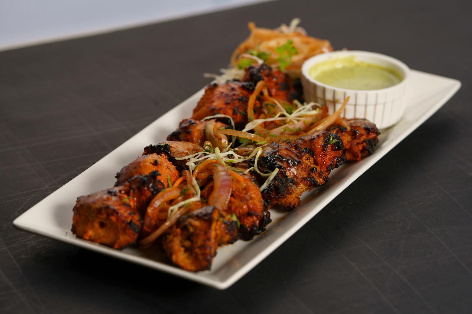 A flavorful chicken tikka dish served with a side of green chutney and garnishes on a white platter.