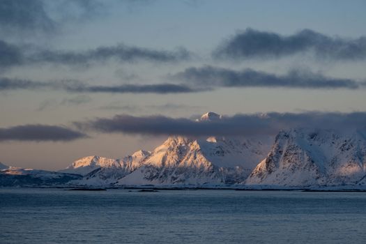 Majestic snow-covered mountains partially obscured by clouds at twilight over a calm sea reflecting serenity.