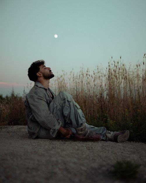 Free A man in casual attire sits on the ground outdoors, gazing at the moon. Stock Photo