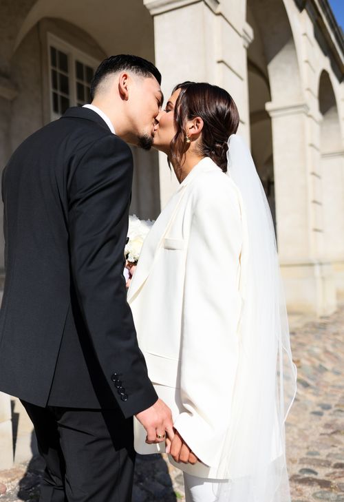 Free A couple shares a romantic kiss in a sunlit courtyard on their wedding day, styled in classic attire. Stock Photo