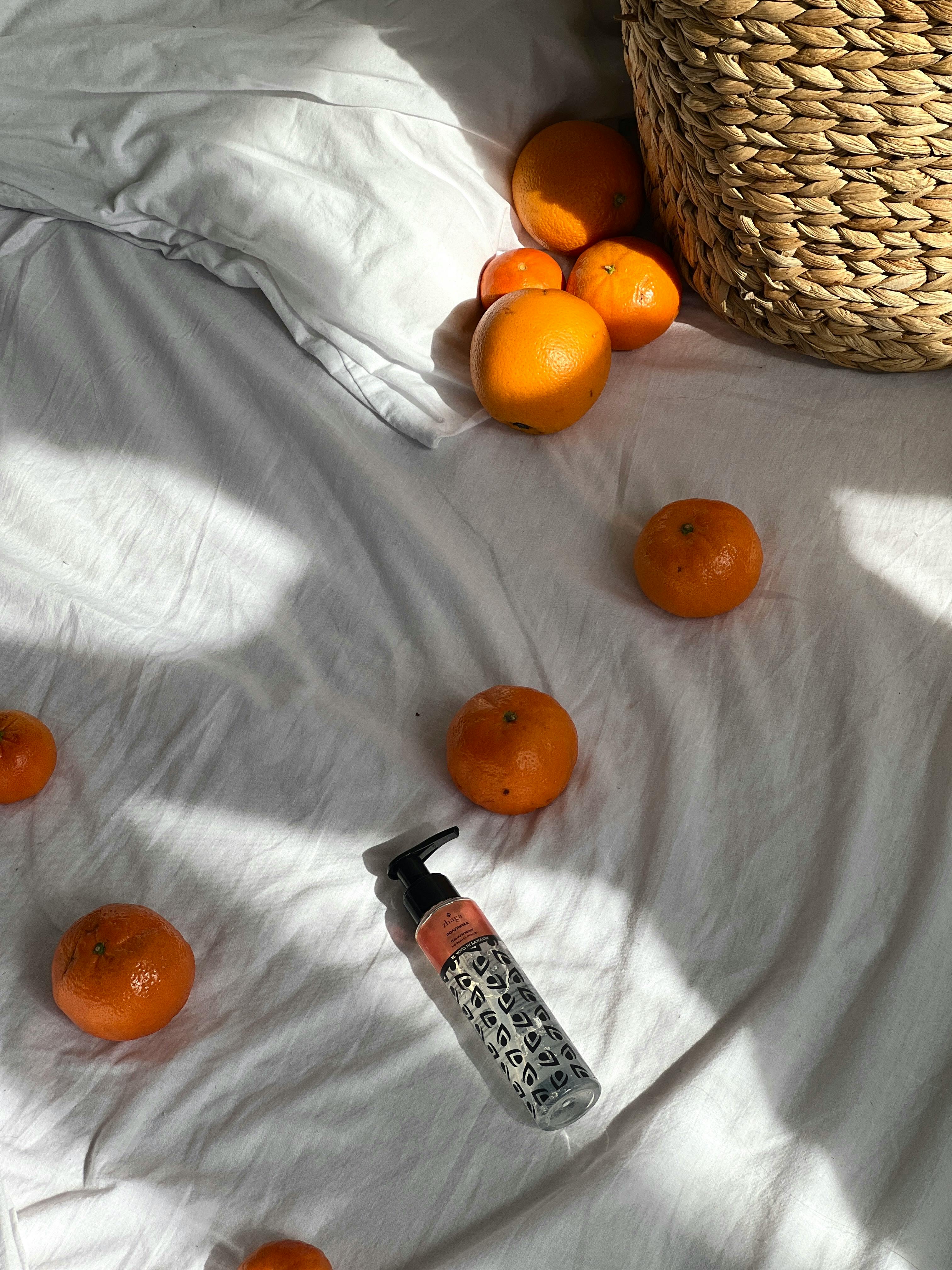 Tangerines and a lotion bottle on a sunlit white bedspread with a woven basket.