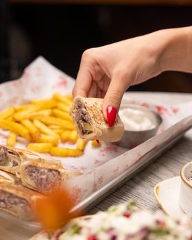 Hand with red nails reaches for a sandwich wrap next to fries, indoor setting