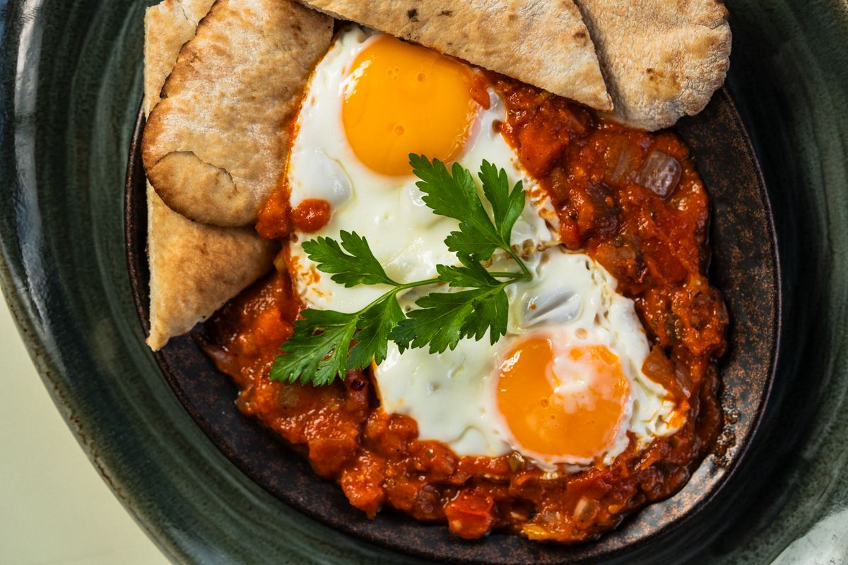 Shakshuka with two runny eggs in chunky tomato sauce, parsley sprig and pita wedges