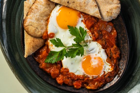 Shakshuka with two runny eggs in chunky tomato sauce, parsley sprig and pita wedges