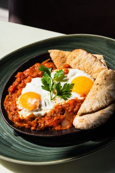 Tasty shakshouka with poached eggs served with pita bread on a green plate.