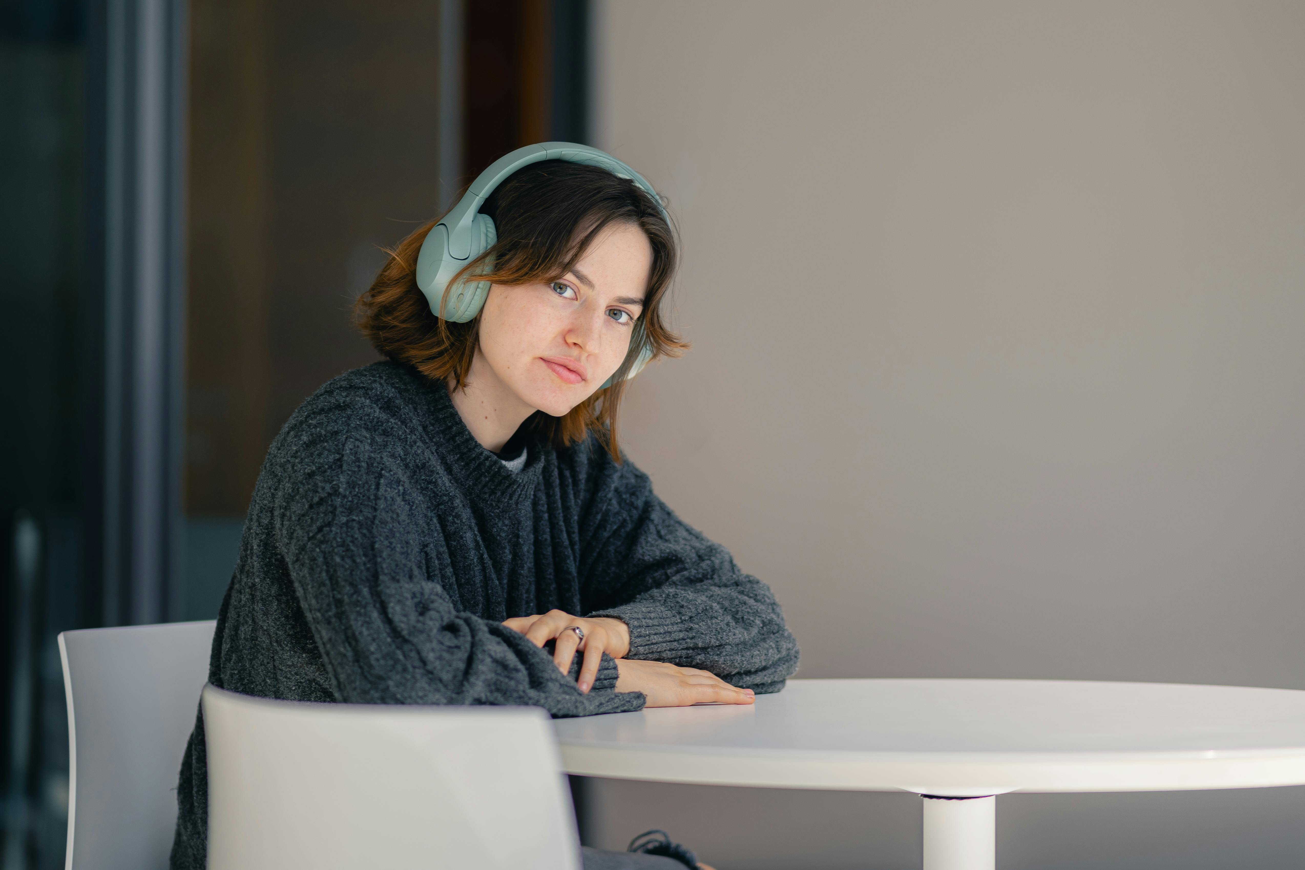 Δωρεάν Μια νεαρή γυναίκα που φοράει ακουστικά κάθεται σκεφτική σε ένα τραπέζι σε εσωτερικό χώρο. Φωτογραφία από στοκ φωτογραφιών