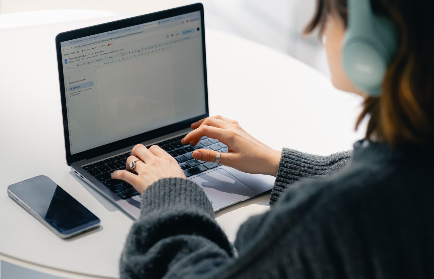 Young woman wearing headphones, typing on a laptop with nearby smartphone on a white table.